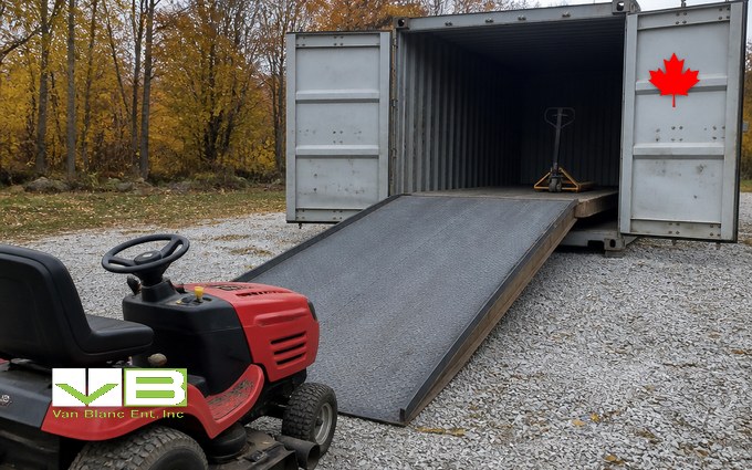 A long steel ramp with a grippy tread surface running up into an open grey container that holds a pallet jack, with a red ride-on mower parked at the foot of the ramp on gravel and autumn trees behind