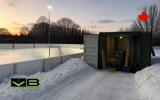 A dark green steel container with its doors open beside a boarded outdoor rink at dusk, holding shovels, a hose reel and spare boards, with snowbanks, a hockey net, a light pole and bare trees