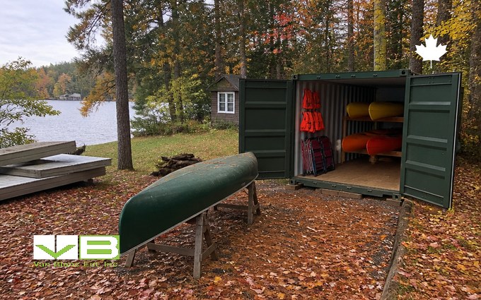 A forest green steel container with its doors open on a leaf-covered cottage lot, holding yellow and red kayaks on racks and orange life jackets on hooks, with a green canoe upturned on sawhorses, stacked dock sections and a small cottage by the lake