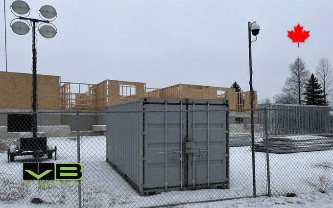 A closed grey steel container with a lock box on its doors inside a chain link fence on a snowy construction site, with a light tower, a camera pole and a partly framed building behind