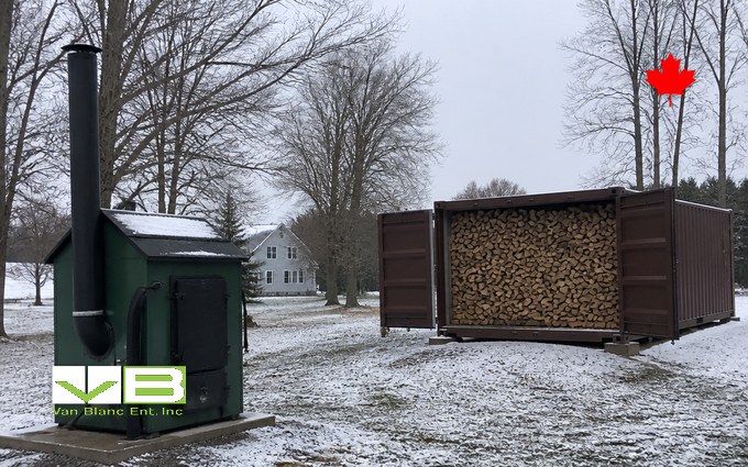 A green outdoor wood furnace with a tall black chimney on a concrete pad in light snow, beside a brown steel container with its doors open and packed with split firewood, bare trees and a farmhouse behind
