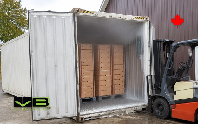 A white refrigerated container with cold mist at its open doors and stacked pallets of plain cardboard produce boxes inside, a forklift parked beside it against a barn wall