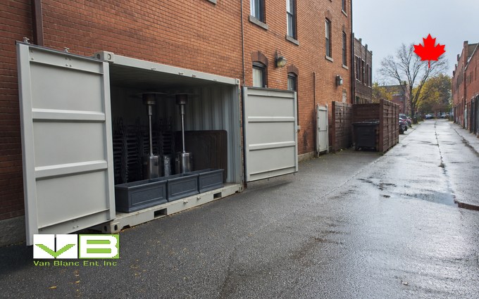 A light grey steel container with its doors open against a brick building in a wet back lane, holding two tall patio heaters, stacked dark bistro chairs and planter boxes, with the lane running past more brick buildings and bare trees