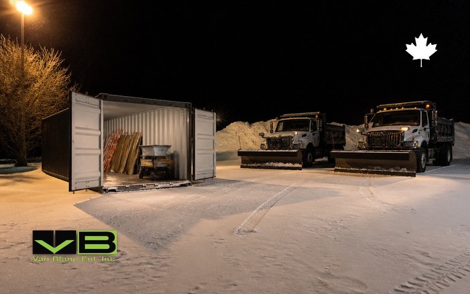 Two plow trucks parked under a yard light on fresh snow beside an open dark grey container holding spare plow blades and a tailgate salt spreader, before dawn