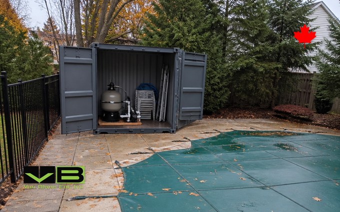 A grey 10 foot steel container with both doors open at the corner of a pool deck, holding a sand filter and pump on a pallet, a stack of white chairs and a folded ladder, beside an in-ground pool under a green mesh safety cover dotted with fallen leaves