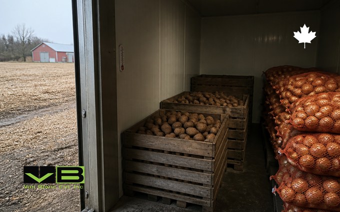 Wooden slatted crates of potatoes and a tall stack of mesh sacks of onions inside a white-walled insulated container, with a harvested field and a red barn through the open door