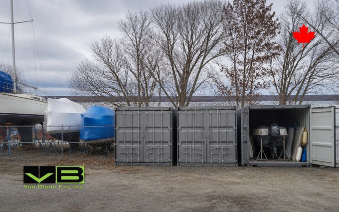 Three grey steel containers in a row along a marina fence, the end one open to show outboard motors on stands, fenders and folded covers, with shrink-wrapped boats on cradles and a lake behind bare trees