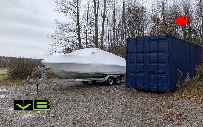 A bowrider boat sealed in tight white shrink wrap on a twin-axle trailer, parked on wet gravel beside a tall dark blue steel container, with bare trees and a grey autumn sky behind