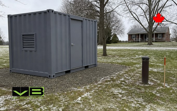 A short grey steel container with a louvered vent and a side man door on a gravel pad in a rural yard dusted with early snow, a few metres from a capped well casing and an orange marker stake, with a farmhouse and bare trees behind