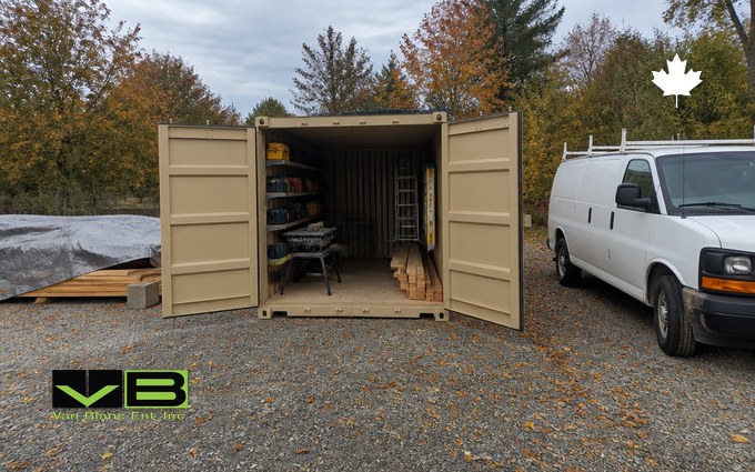 A tan steel container with its doors open on a gravel lot, holding shelves of tool cases, a table saw, a ladder and a stack of lumber, beside a white work van and a tarped lumber pile under autumn trees