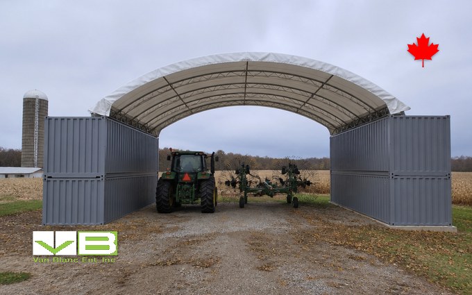 A green tractor and a hay rake parked on gravel under a white fabric arch roof spanning two walls of stacked grey steel containers, with a silo and a harvested corn field behind
