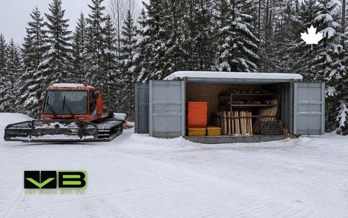 An orange tracked trail groomer parked on a groomed snow lot beside a grey steel container opened wide, holding stacks of blank orange and yellow sign boards, bundles of wooden stakes and shelves of tools, in a snowy spruce forest