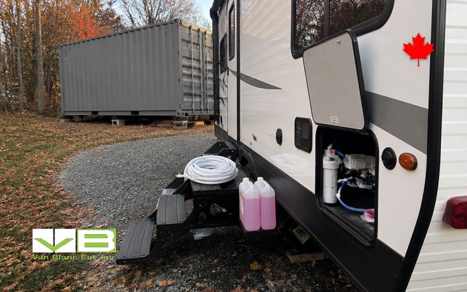A white travel trailer on a gravel pad with its utility hatch open to show the water filter and lines, two jugs of pink antifreeze and a coiled white hose on the entry step, and a long grey steel container on blocks among autumn trees behind