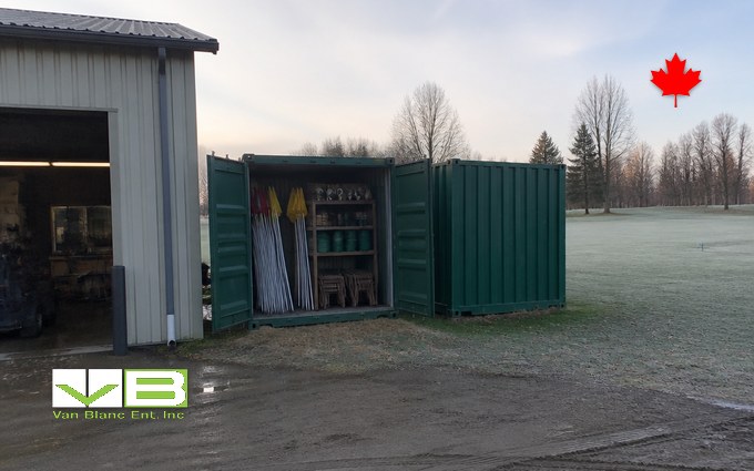 Two green steel containers beside a metal maintenance building on a frosty morning, one open to show bundled flagsticks, tee markers and shelving, with a fairway and bare trees behind