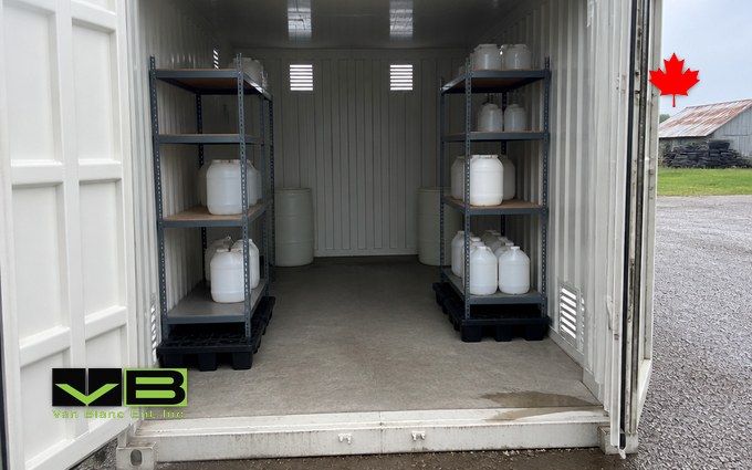 The inside of a white steel container with louvered vents high and low, metal shelving of plain white jugs and drums on black spill containment pallets, and a gravel farmyard through the open doors