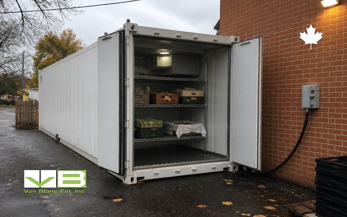 A white refrigerated container with its doors open beside a brick building, its power cable running to an outdoor electrical box on the wall, with wire shelving of produce boxes inside